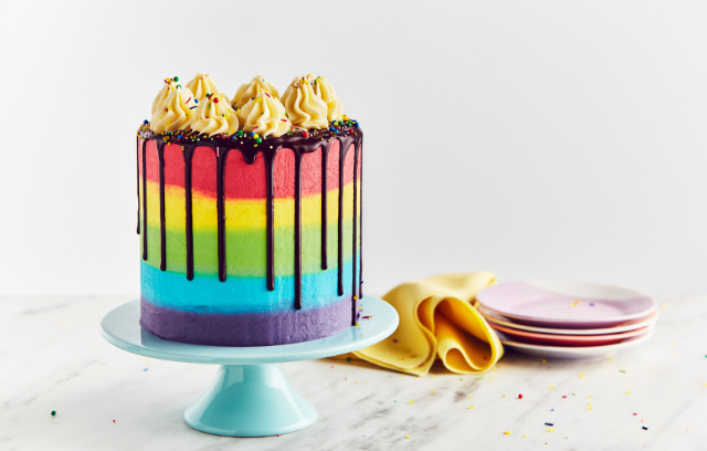 rainbow cake on a cake stand. it is layered with red, then yellow, then green, then blue and then a purple layer of cake. it has chocolate dripping down the sides and is topped with popcorn. in the background of the picture, some plates can be seen. 