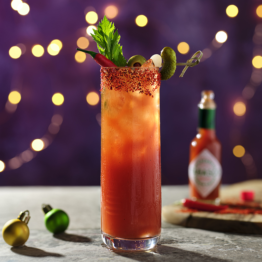 the image shows a red liquid, the bloody mary cocktail, in a tall glass, with crushed chillies around the rim, a celery stick and some tabasco in the background. 